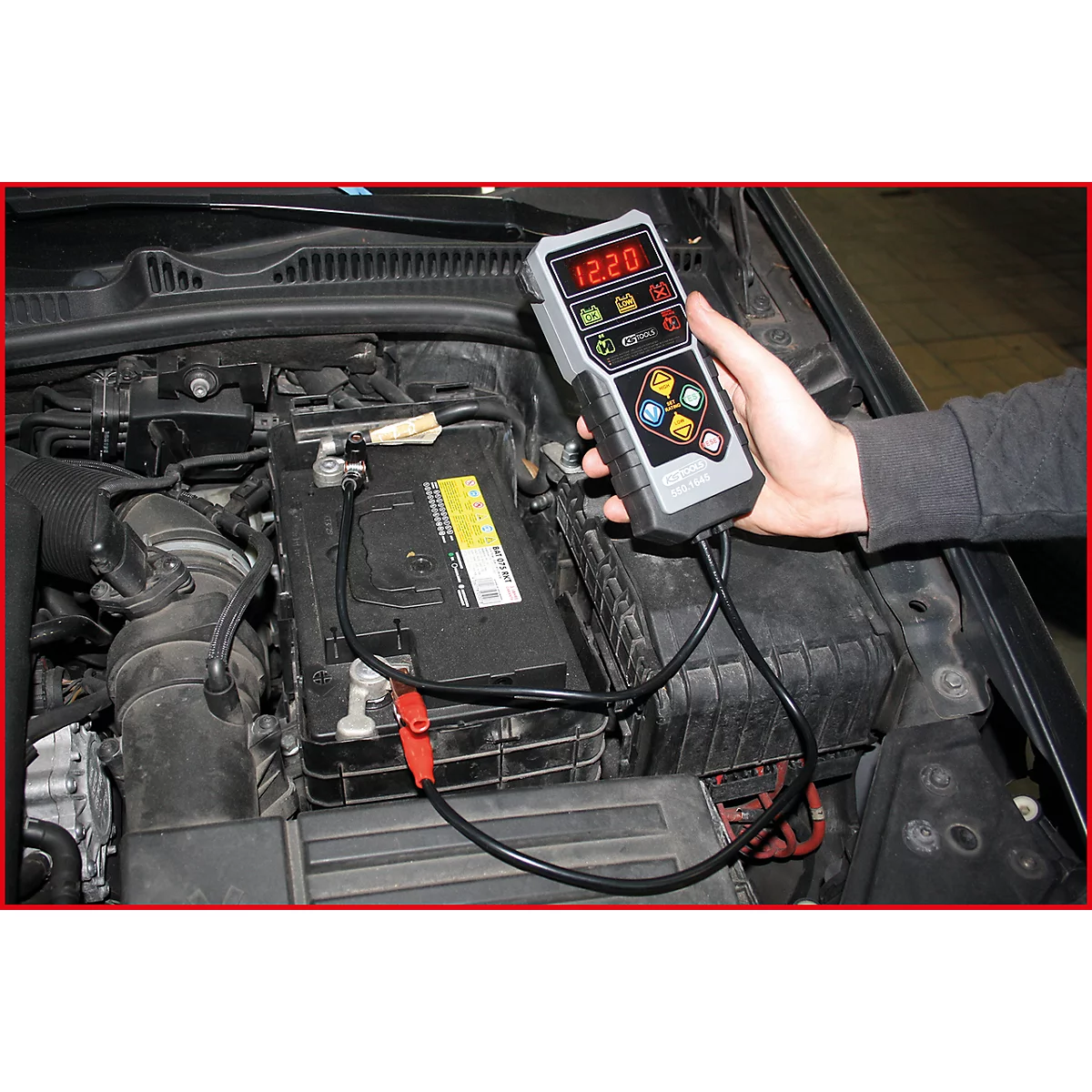 Person misst mit einem Batterietester die Spannung einer Autobatterie im Motorraum. Das Messgerät zeigt 12,20 Volt an.